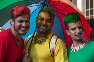 Three people smile beneath a rainbow umbrella. They are dressed in bright colors.