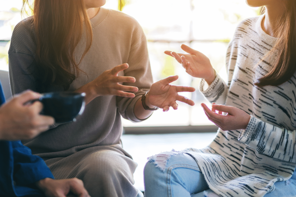 People gesturing in discussion in front of a large, bright window; one person has coffee.