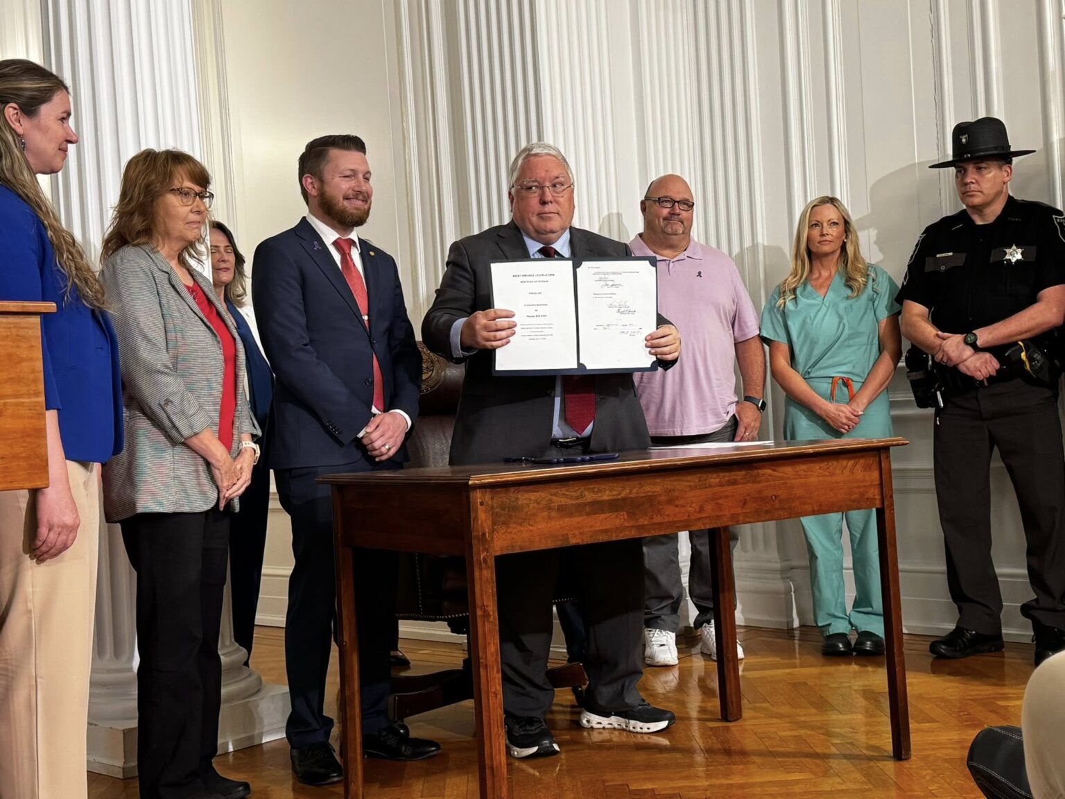 Governor Morrissey stands behind a desk holding up the signed bill, people stand behind him