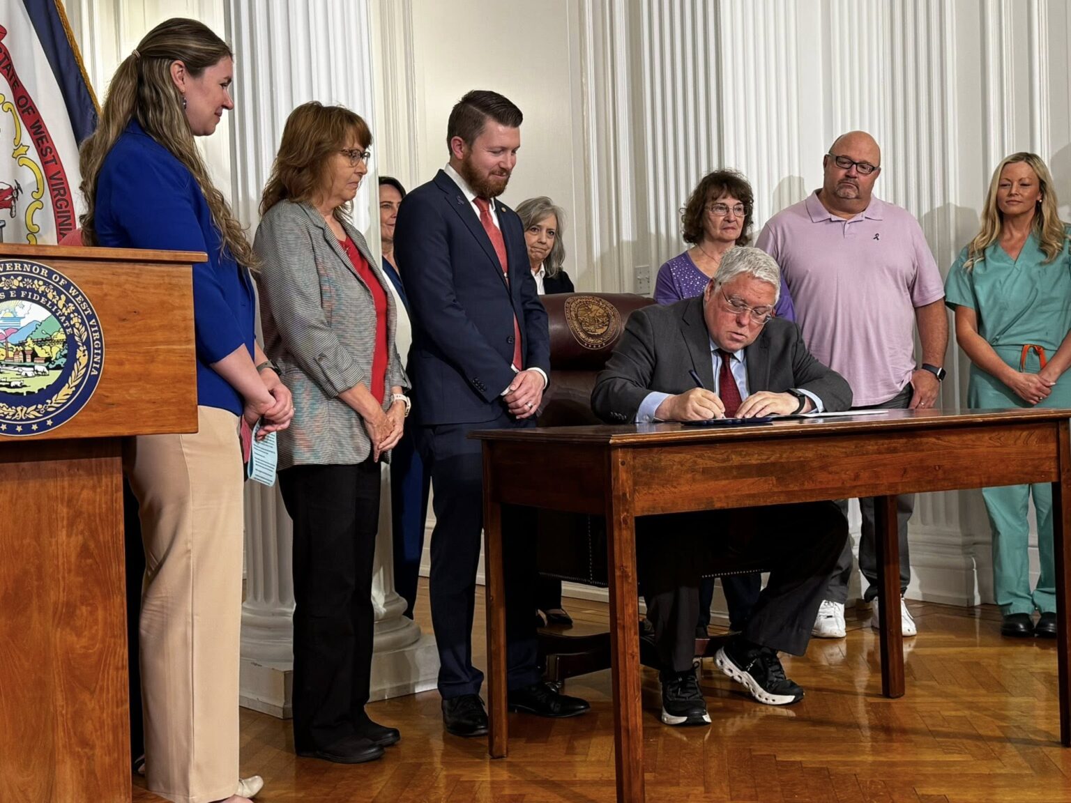 Governor Morrissey sits at a desk signing the bill, people are gathered watching